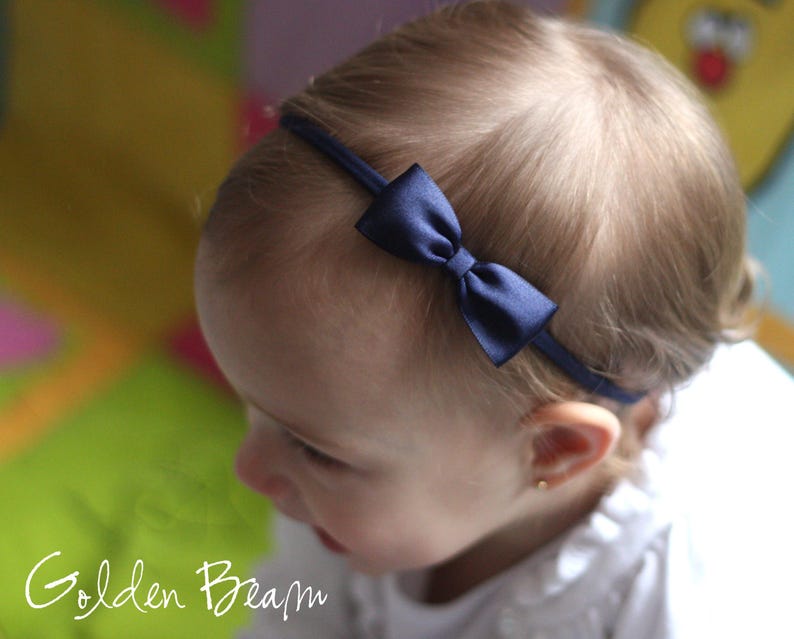 May include: A thin, dark navy blue elastic headband featuring a small, centered satin bow. The accessory is positioned across the top of the head, showcasing a smooth, reflective fabric texture on the bow and a simple, minimalist band design.