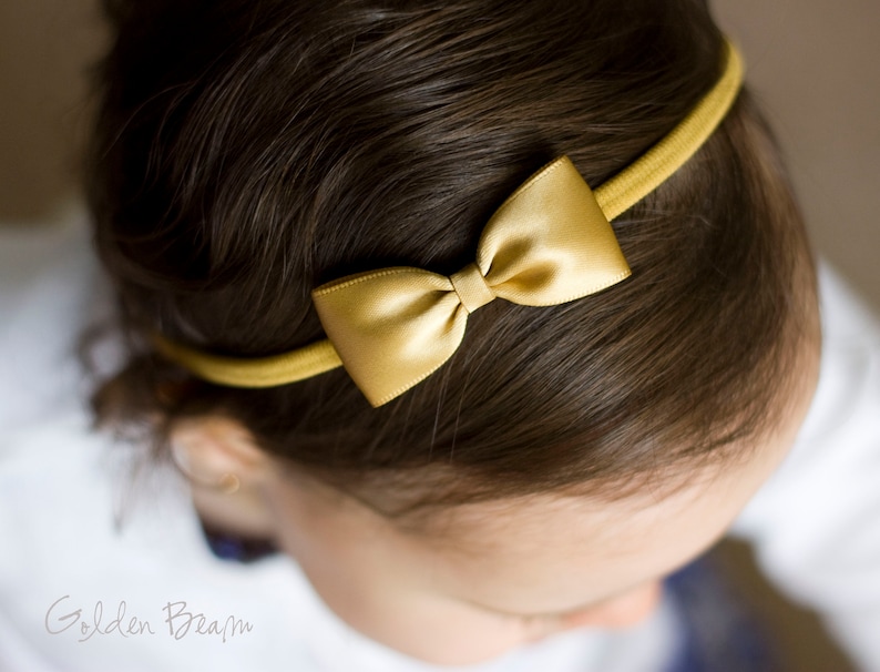 May include: A gold satin bow headband for girls. The headband is made of a soft, stretchy material and is perfect for adding a touch of elegance to any outfit.