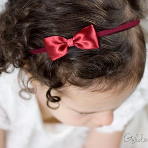 May include: A thin, dark red elastic headband featuring a small, centered satin bow in a matching deep red shade. The accessory is positioned across the top of the head, resting against dark, curly hair. The text Golden Beam is visible.