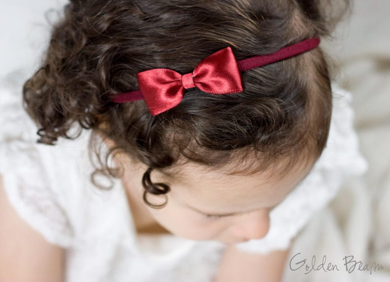May include: A thin, dark red elastic headband featuring a small, centered satin bow in a matching deep red shade. The accessory is positioned across the top of the head, resting against dark, curly hair. The text Golden Beam is visible.