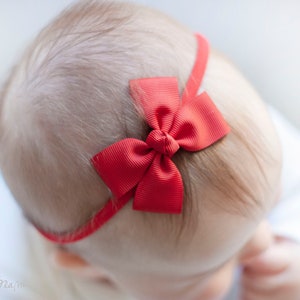 May include: A baby wearing a red bow headband. The headband is made of a red ribbon with a large red bow in the centre.