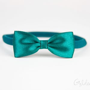 May include: A teal satin bow tie attached to a matching elastic headband. The bow tie features a smooth, shiny finish with a central knot detail. The text Golden Beam is visible in the bottom right corner of the image against a plain background.