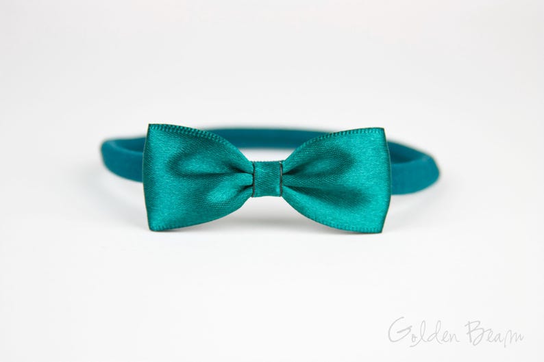 May include: A teal satin bow tie attached to a matching elastic headband. The bow tie features a smooth, shiny finish with a central knot detail. The text Golden Beam is visible in the bottom right corner of the image against a plain background.