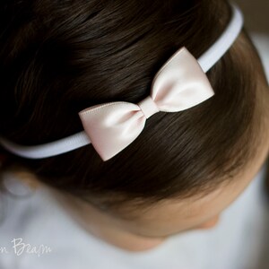May include: A white headband with a light pink satin bow. The headband is worn on a person's head.