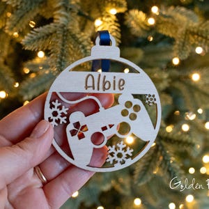 May include: A wooden Christmas bauble with the name "Albie" cut out. The bauble features a video game controller design with snowflake details. A blue ribbon is attached for hanging. The background is a Christmas tree with fairy lights.