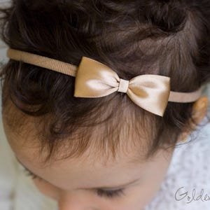 May include: A thin, tan-colored elastic headband featuring a small, centered, metallic gold satin bow. The bow has a smooth, reflective finish and is securely attached to the narrow, ribbed fabric band, which sits across the top of the head.