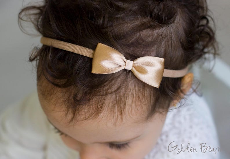May include: A thin, tan-colored elastic headband featuring a small, centered, metallic gold satin bow. The bow has a smooth, reflective finish and is securely attached to the narrow, ribbed fabric band, which sits across the top of the head.