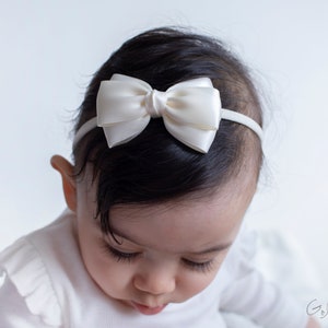 May include: A white satin bow headband for babies and toddlers. The headband is made of soft, stretchy elastic and features a delicate white satin bow.
