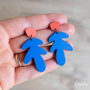 May include: A pair of wooden earrings featuring a blue leaf-like design with a coral-colored top accent. The earrings are connected by a small metal ring. The earrings are held in a hand.