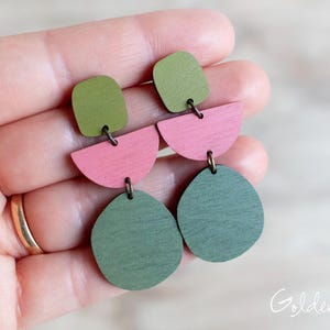 May include: Dangle earrings featuring three stacked geometric wooden shapes in olive green, dusty pink, and dark forest green. The components are connected by small metal jump rings, creating a layered, multi-part drop design.