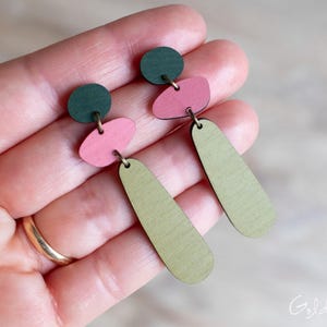 May include: A pair of dangle earrings featuring three stacked geometric shapes in dark green, muted pink, and olive green. The components are connected by small metal jump rings, creating a layered, elongated silhouette with a smooth, matte finish.