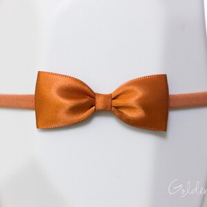 May include: A burnt orange satin bow attached to a matching thin elastic headband. The bow features a smooth, shiny finish and a central knot detail. The text Golden Beam is visible in the bottom right corner of the image.