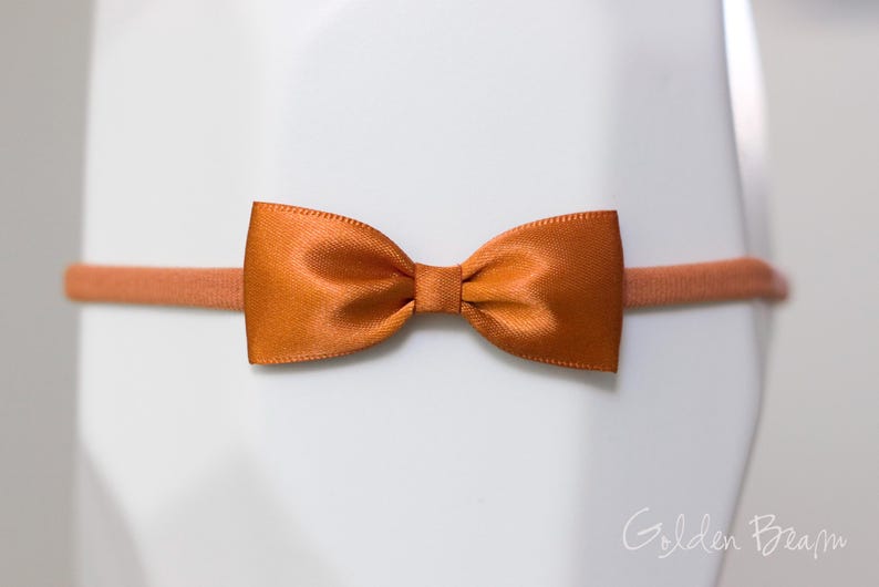 May include: A burnt orange satin bow attached to a matching thin elastic headband. The bow features a smooth, shiny finish and a central knot detail. The text Golden Beam is visible in the bottom right corner of the image.