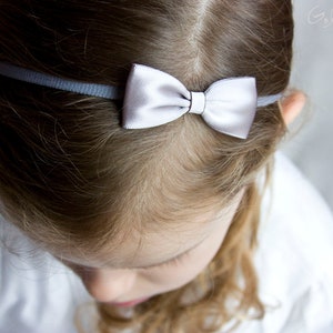 May include: A young girl with blonde hair wearing a gray headband with a white satin bow.