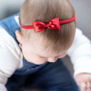 May include: A baby wearing a white shirt and blue overalls with a red bow headband.