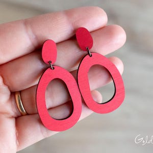 May include: Red wooden earrings with a drop oval design. The earrings have a small, rounded top piece connected to a larger, open oval shape. The earrings are a vibrant red color and appear to be lightweight.