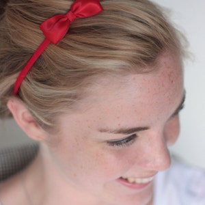 May include: A woman with blonde hair is wearing a red headband with a bow. The headband is thin and has a simple design.