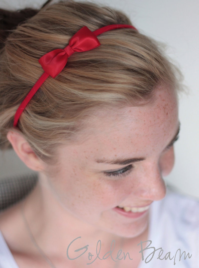 May include: A woman with blonde hair is wearing a red headband with a bow. The headband is thin and has a simple design.