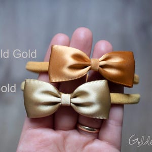 May include: Two satin bow headbands on thin elastic bands. The top bow is labeled Old Gold and the bottom bow is labeled Gold. The bows have a smooth, shiny finish and are centered on the elastic bands. The text Golden Beam is in the corner.