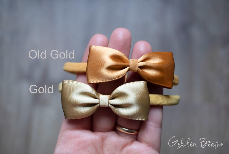 May include: Two satin bow headbands on thin elastic bands. The top bow is labeled Old Gold and the bottom bow is labeled Gold. The bows have a smooth, shiny finish and are centered on the elastic bands. The text Golden Beam is in the corner.