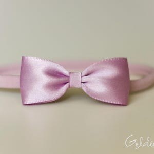 May include: A light pink satin bow attached to a matching thin elastic headband. The bow features a smooth, reflective finish with a central knot detail. The text Golden Beam is visible in the bottom right corner of the image.