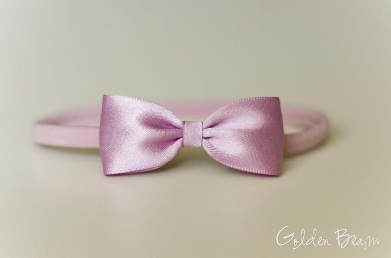May include: A light pink satin bow attached to a matching thin elastic headband. The bow features a smooth, reflective finish with a central knot detail. The text Golden Beam is visible in the bottom right corner of the image.