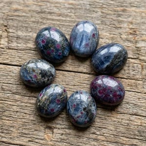 May include: Seven oval-shaped cabochon gemstones arranged in a circular pattern. Each stone features a polished surface with a deep blue and grey base colour, accented by distinct, vibrant magenta and dark red inclusions throughout the material.