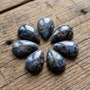 May include: A collection of seven teardrop-shaped cabochons arranged in a circular pattern. Each stone features a polished, mottled surface with a deep blue and grey base color, accented by small, distinct inclusions of vibrant, opaque red mineral deposits.