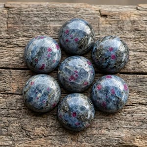Peut inclure: Sept pierres polies de forme sphérique disposées en grappe circulaire. Chaque pierre présente une base marbrée bleue et grise avec des inclusions magenta-rose vibrantes et de petites taches métalliques dispersées sur la surface.