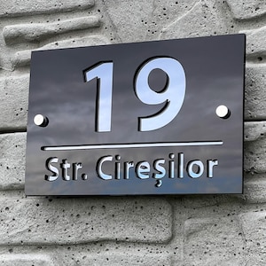 May include: A black rectangular address plaque displaying the number "19" in silver, and the text "Str. Cireșilor" below. The plaque is fixed to a textured grey concrete wall with two silver screws.