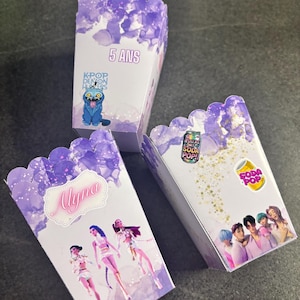 Peut inclure: Trois boîtes à popcorn blanches avec des bords festonnés et des motifs aquarelles violets. Une boîte porte l'inscription "5 ANS" et un chat de dessin animé. Une autre affiche "Alynna" et trois personnages.