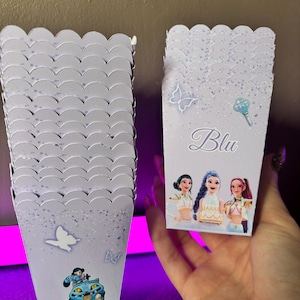 Peut inclure: Plusieurs boîtes à popcorn violet clair avec des bords festonnés. Chaque boîte présente des papillons et des personnages, avec le mot "Blu" en écriture cursive. Une boîte est tenue dans une main.