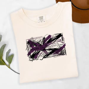 May include: Cream-colored t-shirt with a graphic design. The design features a black and purple abstract shape with the words "Between Structure and Chaos" in a handwritten font. The shirt is folded and has a tag that says "Comfort Colors".