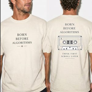 May include: A beige short-sleeve t-shirt featuring black text on the front that reads BORN BEFORE ALGORITHMS. The back displays the same text above a graphic of a cassette tape, with the additional text THINK FIRST SCROLL LATER below it.