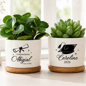 May include: Two white ceramic cylindrical plant pots with bamboo saucers. The left pot features a diploma graphic, the text "Congratulations Abigial" and "Class of 2026". The right pot features a graduation cap graphic, the name "Caroline" and "2026".