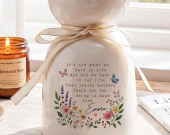Könnte beinhalten: Eine weiße Keramikvase mit gewelltem Rand, gefüllt mit rosa und weißen Alstroemeria-Blüten. Ein cremefarbenes Band ist um den Hals gebunden. Die Vase zeigt ein Blumenmuster, Schmetterlinge und den Text: It's not what we have in life, but who we have in our life, that really matters. Thank you for being in mine. Love, Emma.