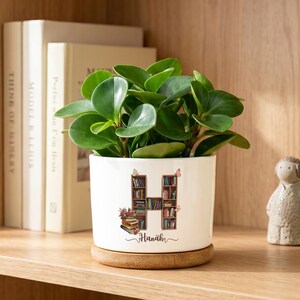 May include: A white ceramic plant pot with a bamboo saucer containing a green Peperomia plant. The pot features a graphic of the letter H shaped like a bookshelf filled with books, small butterflies, and the name Hanah printed in black script.
