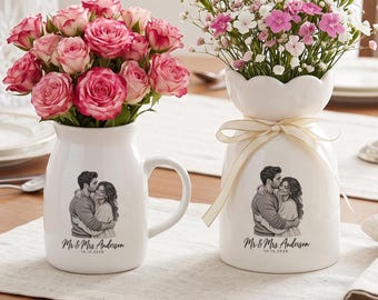 Op de afbeelding: Twee witte keramische vaten, een mok en een vaas, elk voorzien van een zwart-wit illustratie van een omhelzend stel met de tekst Mr & Mrs. Anderson 10.15.2026. De mok bevat roze rozen, terwijl de vaas roze en witte bloemen bevat.