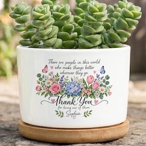 Puede incluir: Una maceta de cerámica blanca con base de madera que contiene una suculenta verde. La maceta presenta un gráfico floral con mariposas y el texto: There are people in this world who make things better wherever they go. Thank You for being one of them. Sophia.