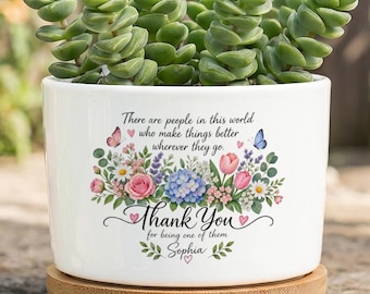 May include: A white ceramic planter with a wooden base containing a green succulent. The planter features a floral graphic with butterflies and the text: There are people in this world who make things better wherever they go. Thank You for being one of them. Sophia.