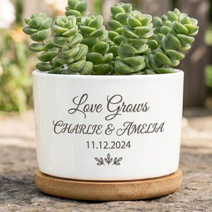 May include: A white ceramic cylindrical planter containing a green succulent plant. The planter features the black printed text "Love Grows", "CHARLIE & AMELIA", and "11.12.2024" with a small leaf icon. It sits on a circular light brown wooden base.