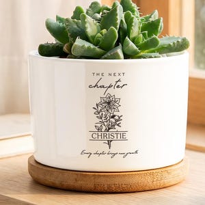 Puede incluir: Una maceta cilíndrica de cerámica blanca que contiene una suculenta verde. La maceta presenta texto en negro que dice THE NEXT CHAPTER, CHRISTIE y Every chapter brings new growth, junto con una ilustración floral sobre una base de madera.