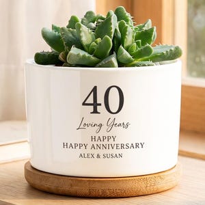 May include: A white ceramic cylindrical planter with a bamboo saucer containing a green succulent. The planter features black text that reads 40 Loving Years, HAPPY HAPPY ANNIVERSARY, and the names ALEX & SUSAN printed on the front surface.