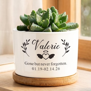 May include: A white ceramic cylindrical planter containing a green succulent. The planter features black text reading Valerie, a heart with wings and a halo, and the phrase Gone but never forgotten. 01.19-02.14.26. It sits on a round wooden base.
