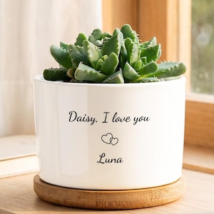 Puede incluir: Una maceta cilíndrica de cerámica blanca que contiene una planta suculenta verde. La maceta presenta el texto impreso en negro "Daisy, I love you" sobre dos pequeños corazones y el nombre "Luna", sobre una base circular de madera.