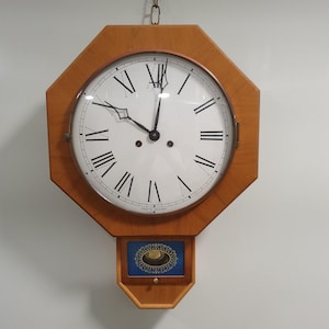 May include: A wooden wall clock with a light brown octagonal frame and a white face. The clock features black Roman numerals, black hands, and a small blue and gold decorative element at the bottom. The clock is suspended from a chain.