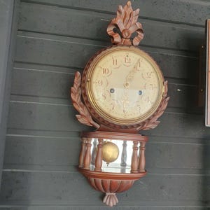 Puede incluir: Un reloj de pared de madera vintage con una esfera redonda y números romanos. El reloj tiene un marco decorativo de madera tallada y un péndulo. El reloj está montado en una pared gris oscuro.