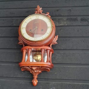 May include: A decorative wooden wall clock with a round face and Roman numerals. The clock features a pendulum and is encased in a reddish-brown wooden frame. The clock is mounted on a dark, textured wall.