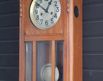 Op de afbeelding: Een houten wandklok met een lichtbruine afwerking. De wijzerplaat is rond met zwarte cijfers en wijzers. Een glazen slinger zwaait eronder. De klok is rechthoekig met decoratieve details.