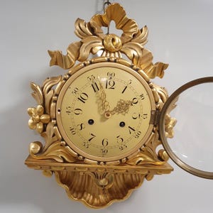 Puede incluir: Un reloj de pared dorado con un diseño ornamentado. La esfera del reloj es de color crema con números y manecillas negras. El reloj tiene un lazo decorativo y detalles florales. La cubierta de vidrio del reloj está abierta.